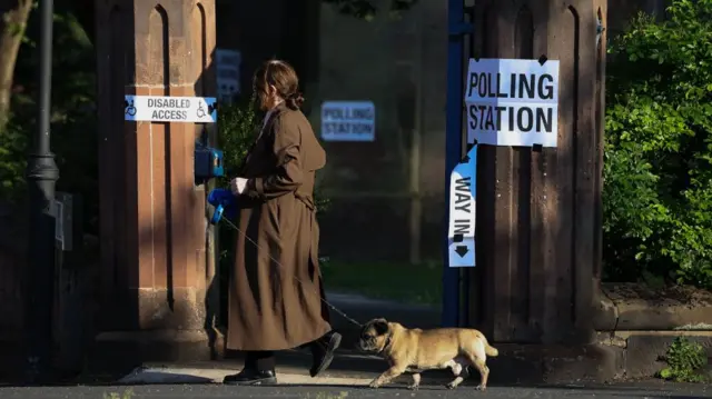 A woman with a small dog on a lead walk past a building with a sign saying Polling station.