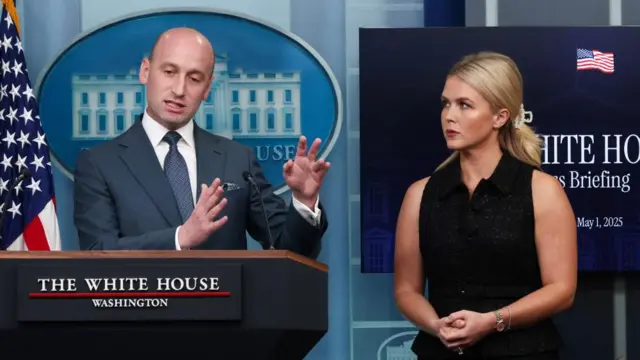 US White House Deputy Chief of Staff Stephen Miller speaks next to White House Press Secretary Karoline Leavitt during a press briefing. Miller wears a suit and white shirt, while Leavitt wears a black sleeveless top. Miller stands behind a podium.