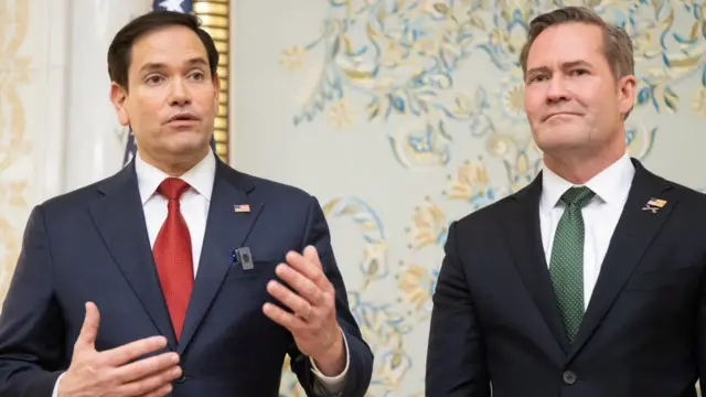 Marco Rubio stands on the left, wearing a suit and red tie. Mike Waltz stands on the right, wearing a suit and green tie. Rubio is speaking.