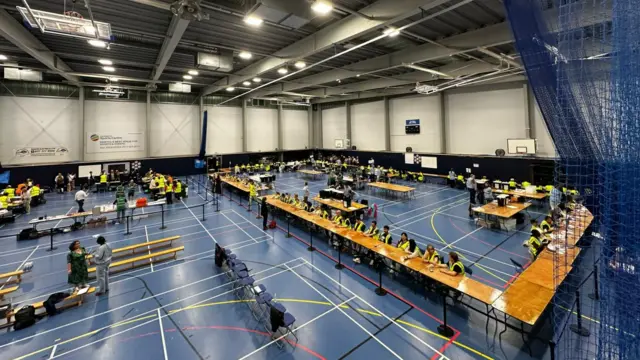 View from above on rows of tables where people are counting votes. It is a sports hall with a blue floor.