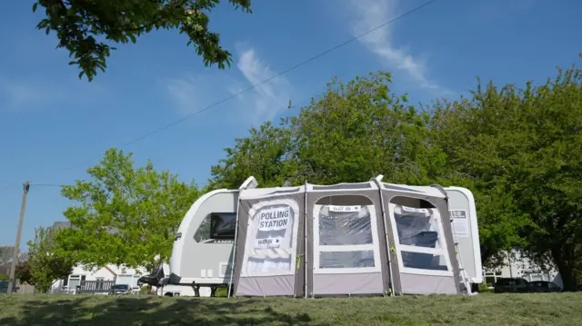 A caravan stands in a field, a canvas stretches over it with the sign 'Polling Station'