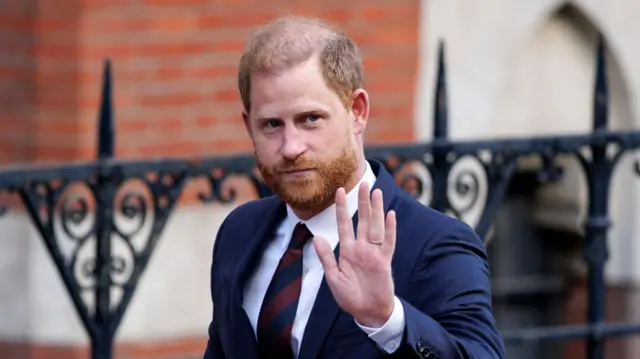 Prince Harry with his left arm raised as he walks along street. He's wearing a navy suit, white shirt and navy and maroon striped tie