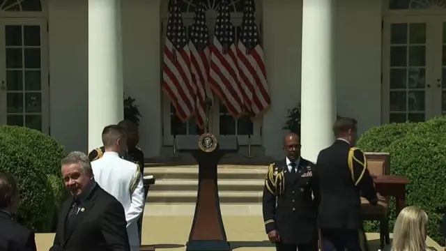 In a garden in front of doors and steps, a podium stands in the middle with a microphone on it. Service men stand about in front