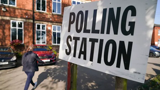A woman (L) in black jacket, jeans and white trainers walks through a parking lot headed for a red-bricked building. In the foreground is a white sign reading Polling Station