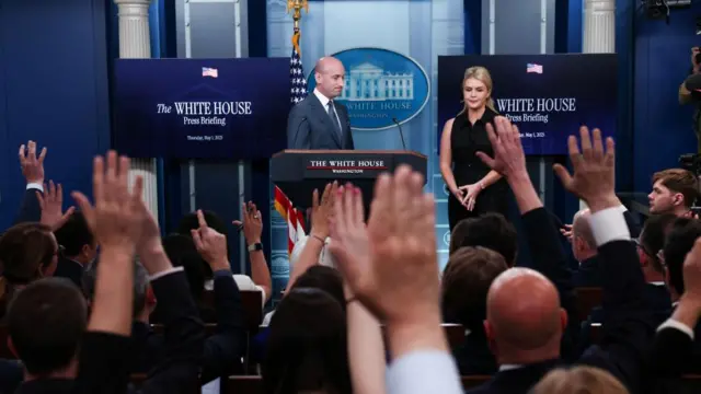 U.S. White House Deputy Chief of Staff Stephen Miller and White House Press Secretary Karoline Leavitt stand at a press briefing with a crowd of reporters in front of them, many with their hands up to ask a question.
