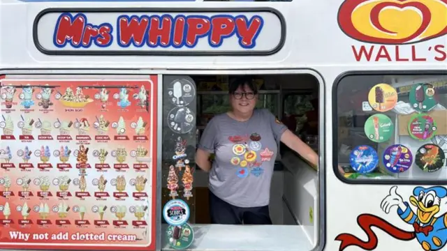 A woman in an ice cream van with Mrs Whippy written above the serving hatch, to the left is a board showing pictures of the ice creams on sale.