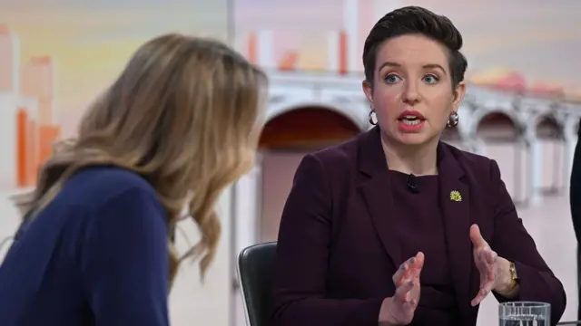 Carla Denyer is sitting at a table speaking to the camera with her hands slightly raised. She has short, dark hair and is wearing a purple suit.