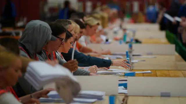 People wearing orange vests sit at a long row of tables counting through election results