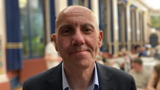 Rob Waltham smiles to camera at the count. He is wearing a suit without a tie