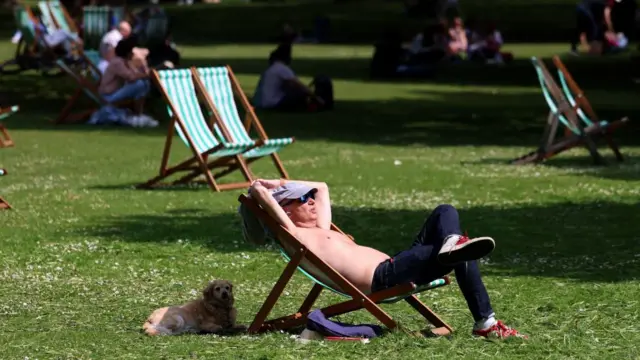 A man sunbathes shirtless in a deck chair in a park, a dog is sheltering in the shade of the chair behind him
