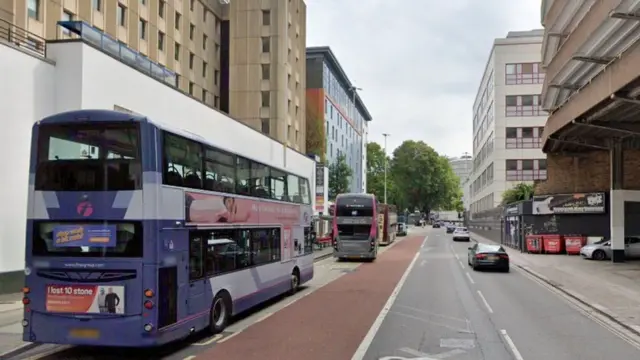 A line of buses waiting at a bus stop in Bristol city centre on the left. There are tall buildings on either side of the road, which has a few cars on it.