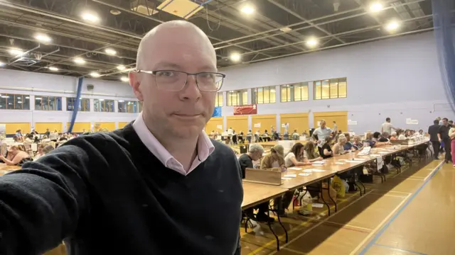 A selfie taken by Martin Jones. He is wearing a jumper over a shirt. Behind him are rows of tables with people counting votes.