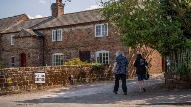 A man (L) with long white and grey hair walks hand in hand with a woman (R) with long brown hair towards a polling station hosted inside a red-bricked building with white windows