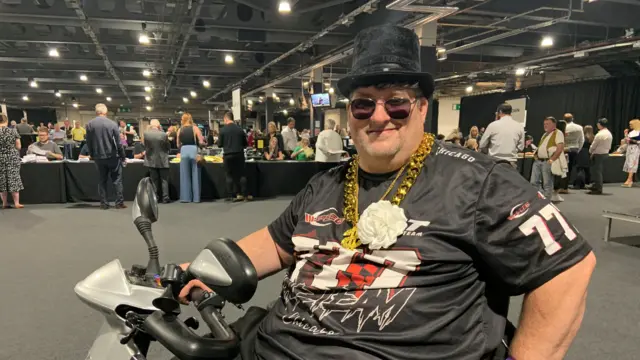 Richie Vallance wearing a black t-shirt and top hat, and a white rosette