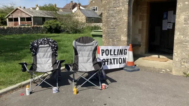 Two grey and black camping chairs placed at the entrance of an old building (seemingly a church) with a sign reading polling station held up between two traffic cones, some low houses in the background