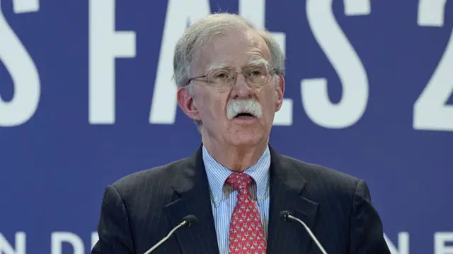 Bolton, in a striped suit, speaks into two mics with a blue background behind him.