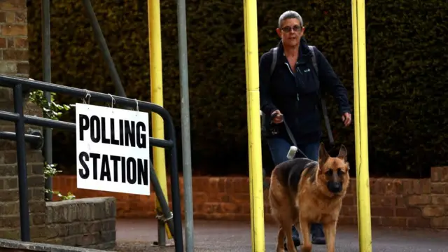 Woman and Alsation dog walking towards camera with a sign reading polling station.