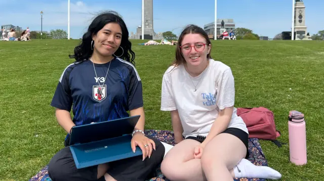 Two women sat on on a floral blanket on some grass, one has a laptop and there is a pink bottle next to her