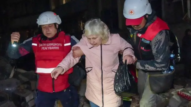 Two emergency service workers help a lady walk over rubble. The image was taken at night - the sky is black