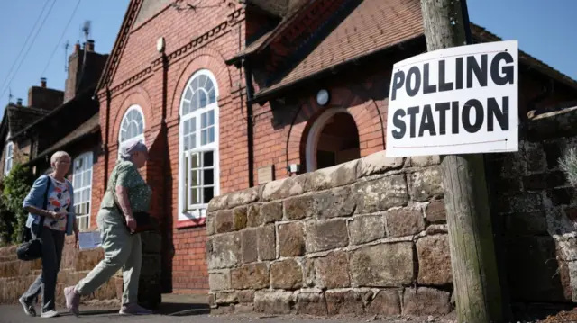 Two women in shirts and trousers, one with a blue cardigan (L) approach a red-bricked building serving as a polling station, a sign marking its designation affixed to a wooden electricity pole to the right