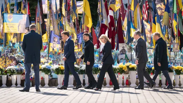 Merz, Macron, President Zelensky, First Lady Olena Zelenska, Starmer, and Tusk walk in front of a memorial in Kyiv's Independence Square, which has lots of flags and flowers. They are all suits.