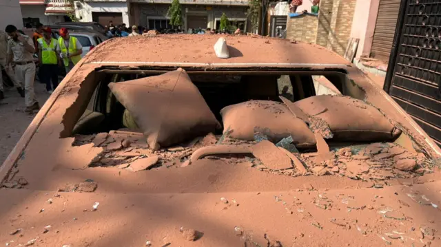A car covered in dust, windscreen broken and people stood behind.