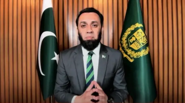 Pakistan's information minister sits between two flags while looking at the camera.