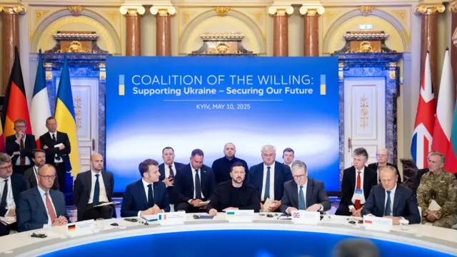 Merz, Macron, Zelensky, Starmer, and Tusk sit at a long, curved desk with officials sat behind them. They is a board up behind them that says Coalition of the Willing: Supporting Ukraine - Securing Our Future.
