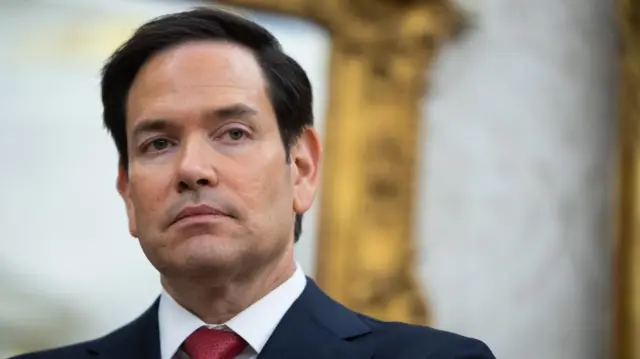 Marco Rubio in a blue suit and red tie, looking at the camera. Background blurred.
