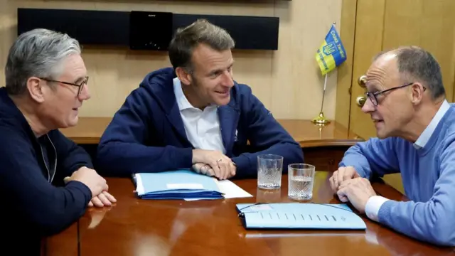 Starmer, Macron and Merz sat around a wooden table on a train. All have arms folded on front of them on the table.