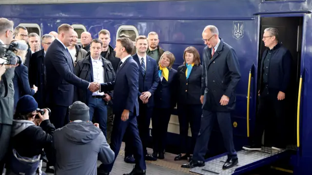 Macron, Merz, and Starmer step off a dark blue train and is greeted by officials on the train station platform.