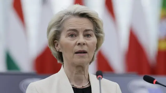 Ursula von der Leyen, European Commission President, speaks behind microphones wearing a white blazer.