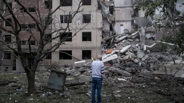 A man standing in front of a building collapsed into rubble in Ukraine.