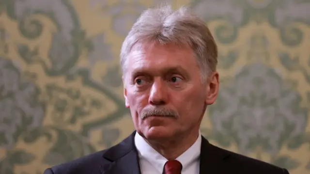 Close up of Dmitry Peskov (middle aged man with short grey hair and moustache) in a black suit, white shirt and red tie stands in front of a wall with patterned wallpaper