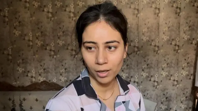 A woman called Tanya stood in a bedroom, wearing a shirt with geometric pattern looking at the camera.