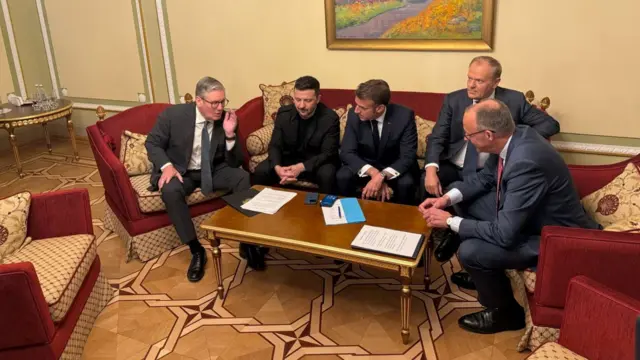 Starmer, Zelensky, Macron, Tusk, and Merz sit on a red sofa and chairs with paperwork on a wooden table. They are talking to each other.