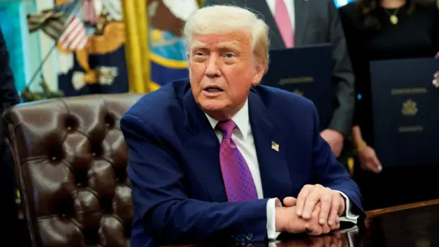 Donald Trump sat in Oval Office with his arms crossed on a table, people stand behind him.