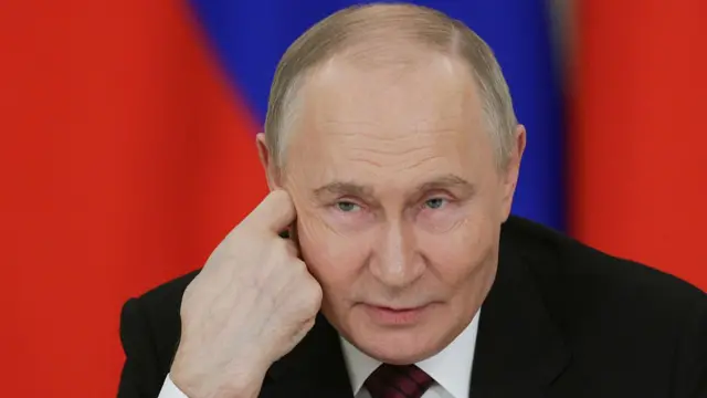 Close up of Vladimir Putin in a black suit, white shirt and burgundy tie with black stipes, two Russian flags behind him, blurred