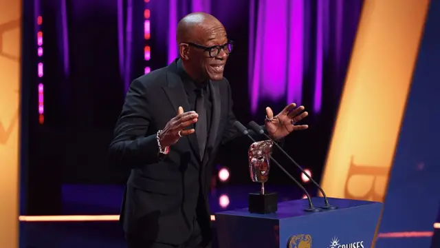 Lennie James with his hands raised as he delivers a speech following his win. He's in an all-black ensemble, his golden Bafta award on the lectern in front of him