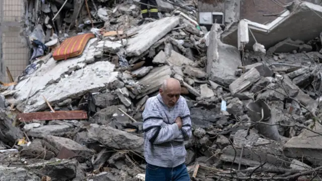 A man stands with arms folded, behind him is a huge pile of rubble and debris in Ukraine.