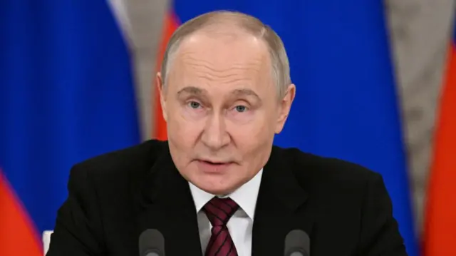 Vladimir Putin in black suit, white shirt and burgundy tie with black stripes sitting in front of two mic. Two Russian flags behind him