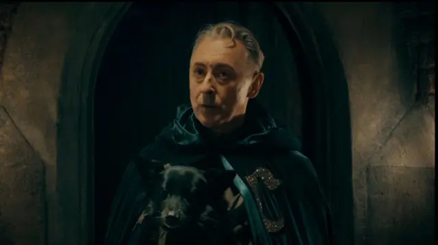 Alan Cumming in a spoof skit of the traitors wearing a green cape and holding a black dog