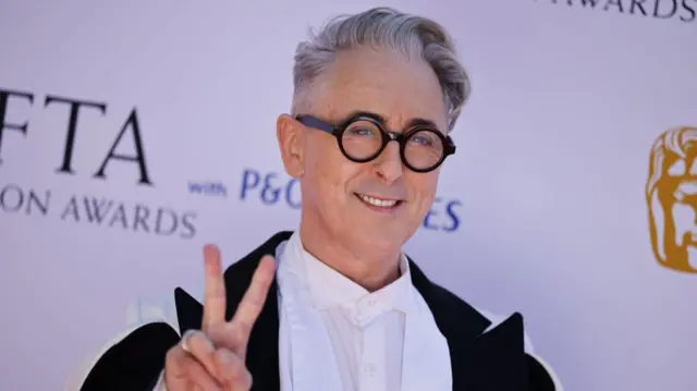Alan Cumming in a black and white evening suit holds his right hand up in a victory sign