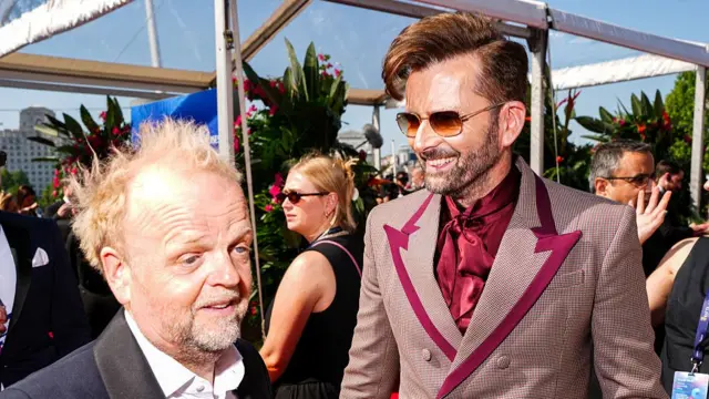 Toby Jones (L) stands next to David Tennant (R) both are in suits, one dark blue and black, the other is red
