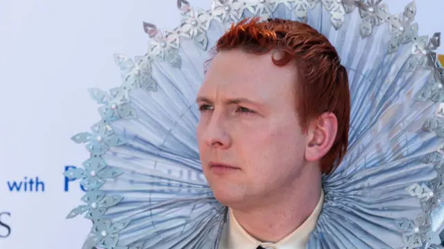 Joe Lycett with red hair on red carpet at 2024 Bafta TV awards in white Elizabethan-inspired collar