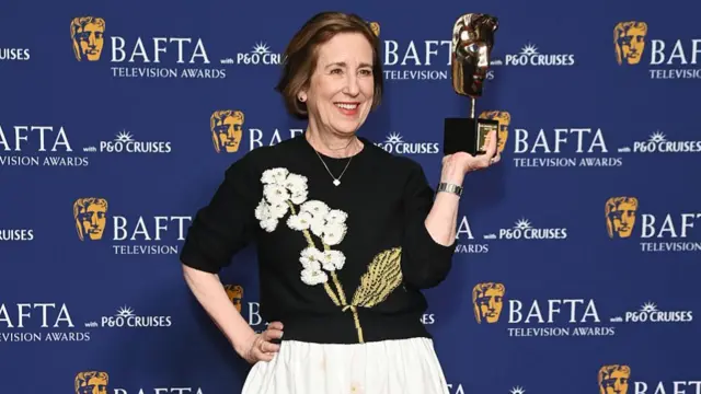 Kirsty Wark in black jumper with white flower decoration and white skirt holds up a Bafta with her left hand