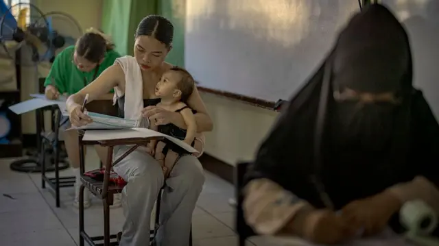 A  young mother wearing a black tube top and long pants, sitting on a chair in a classroom. She is holding a baby on her lap with one hand and holding a pen and paper with the other as she writes down her vote