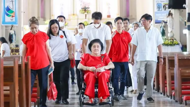 Members of the Marcos's, either wearing red or white shirts, march down the aisle of a church. Imelda Marcos, in a full red attire and puffy black hair, is being pushed on a wheelchair by a man in white.