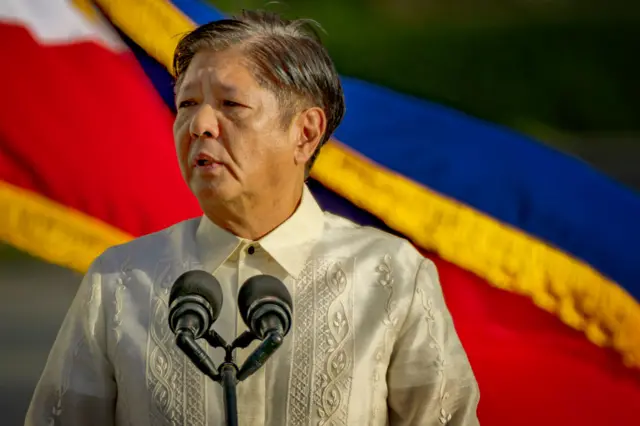 Ferdinand "Bongbong" Marcos Jr wearing a white linen shirt addressing the crowd through a microphone, in front of the Philippine flag