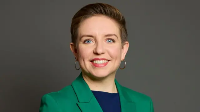 Headshot of Denyer smiling at the camera in front of a grey background